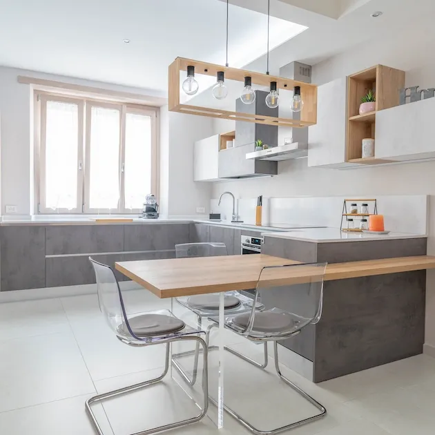 Interni moderni del progetto 'Casa Santa Fe' con cucina angolare in finitura grigio cemento e bianco. In primo piano, un tavolo snack in legno rovere sospeso con sedie trasparenti in policarbonato e lampadario minimalista in legno con lampadine a vista.