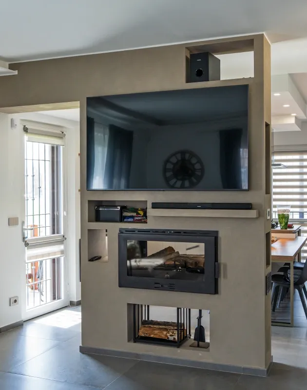 Vista d'interni di un open space moderno con una parete multifunzionale divisoria in microcemento beige. La struttura integra un grande televisore a schermo piatto, un camino a legna moderno ad incasso con vetro, e vani aperti per legna, soundbar e altri accessori. Sullo sfondo si intravedono la zona pranzo e una porta-finestra luminosa che d&agrave; sull'esterno.