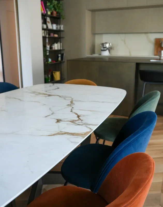Dettaglio della zona giorno nel progetto 'Casa Dani': tavolo da pranzo in marmo bianco con venature oro, circondato da sedie in velluto colorato (ottanio, senape e ruggine). Sullo sfondo, una cucina moderna minimalista con isola e libreria a tutta altezza.